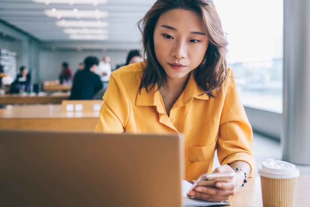 Focused woman looking at laptop screen and using smartphone in workspace in Hong Kong city with people in background